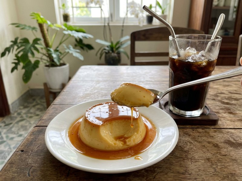 Thìa nhỏ múc một miếng bánh flan caramel mềm mượt tan chảy bên cạnh ly cà phê đen đá mát lạnh